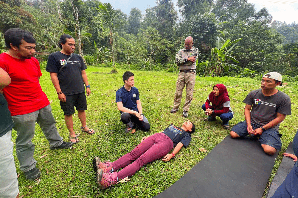 Participants engaged in a Wilderness First Aid course with Viristar in West Java, Indonesia. The scene shows individuals practicing first aid techniques in a natural outdoor setting, surrounded by lush greenery.