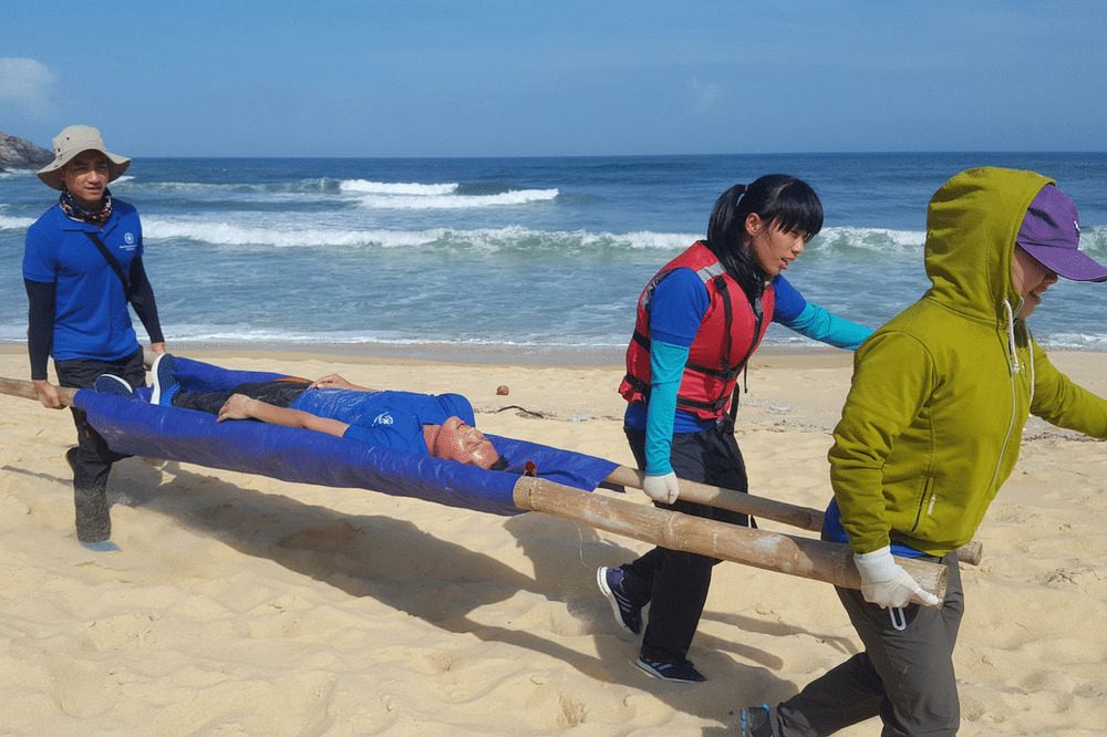 Participants in a Wilderness First Aid course on a sandy beach, demonstrating rescue techniques as they carry a person on a gurney.