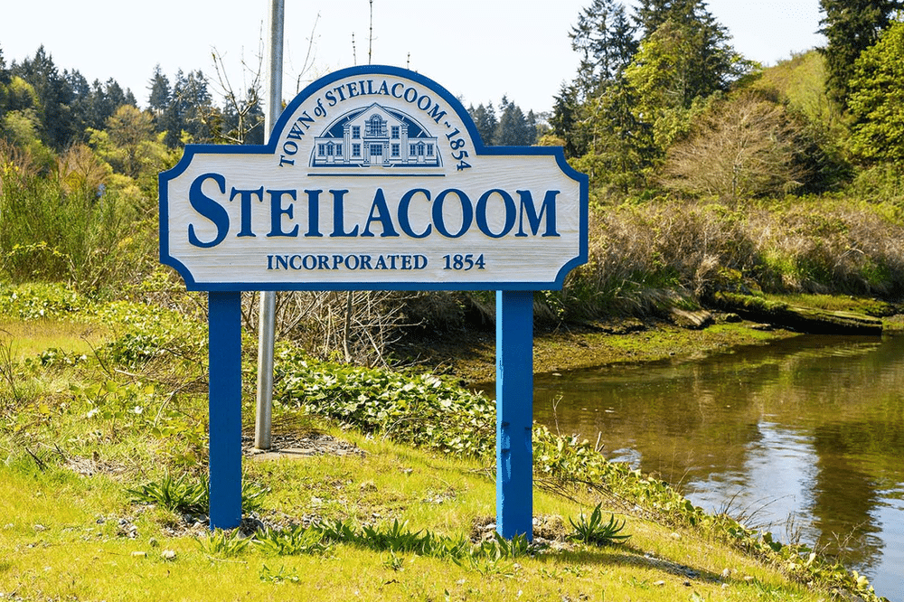 Welcome sign for the town of Steilacoom, featuring the town's name in bold letters surrounded by a decorative border. The sign is set against a backdrop of lush greenery and trees.