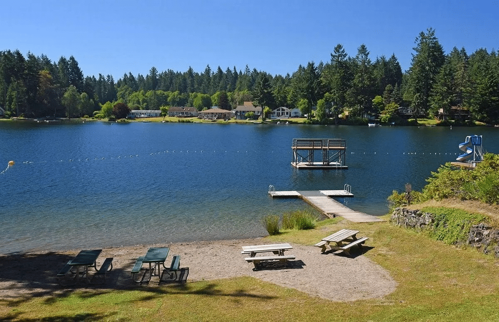 Peaceful view of Florence Lake, also known as the Old Swimming Hole, at Lowell Johnson Park on Anderson Island, Washington. The image features calm waters reflecting surrounding trees, with a sandy beach area in the foreground and lush greenery framing the serene landscape under a clear blue sky.