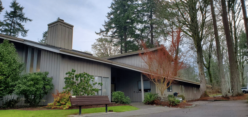 Exterior view of the Community Center run by the town of Steilacoom, showcasing a welcoming entrance with a bench, large windows, and landscaped grounds.