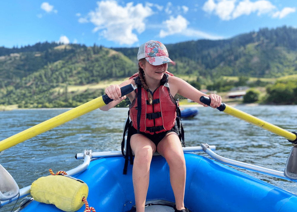 A young girl paddling on a blue raft in a river, with paddles in hand. She is surrounded by lush green mountains in the background under a clear sky, showcasing a serene outdoor adventure.