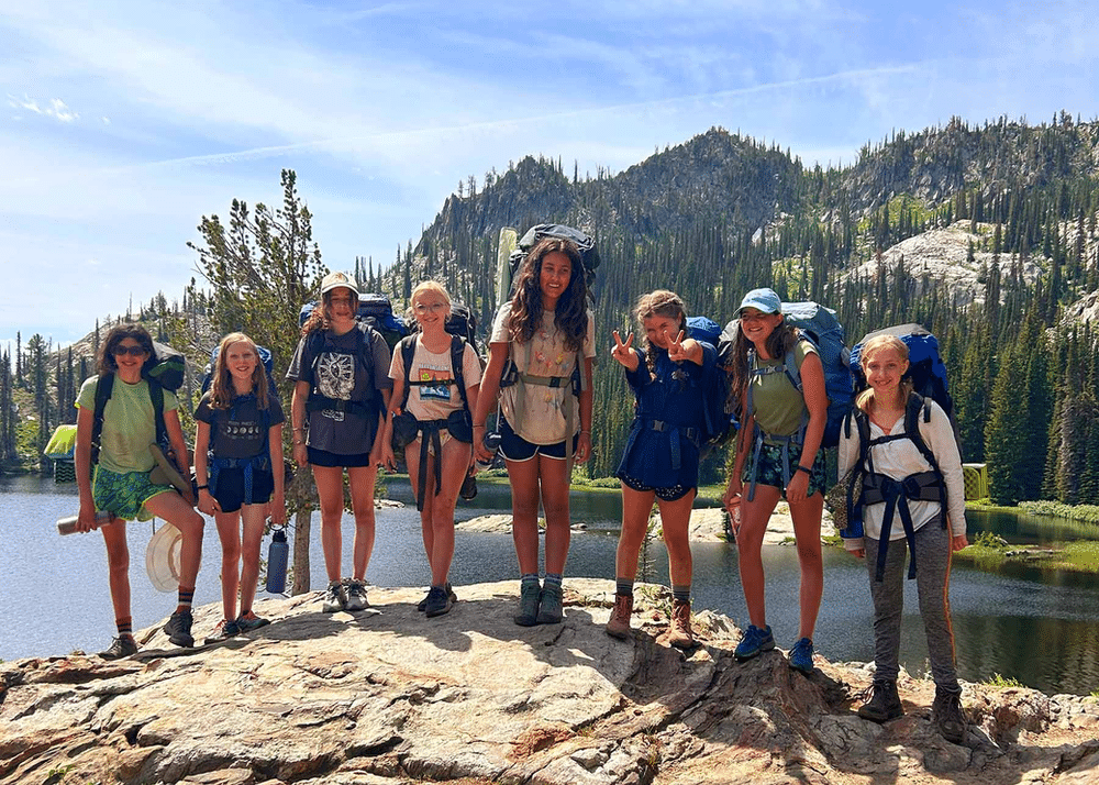 A group photo of Wild Hearts Idaho participants on a backpacking trip, smiling and posing together on a scenic trail. They are dressed in outdoor gear, with backpacks, and surrounded by lush green forests and towering mountains in the background.