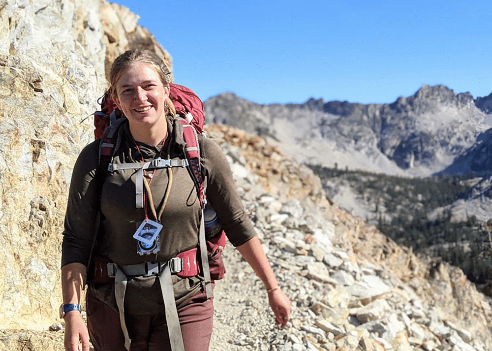 Serena Rasmussen, Executive Director of Wild Hearts Idaho, pictured backpacking through a scenic wilderness area. She is seen wearing hiking gear, a backpack, and surrounded by lush greenery and rugged mountain terrain.