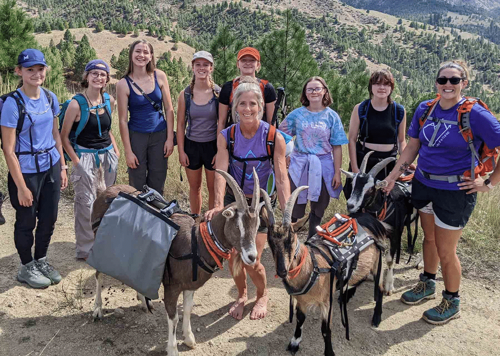 A group of Wild Heart Idaho participants on a goat hike, smiling and posing together outdoors. Two goats, each wearing a backpack, are also featured in the photo, standing alongside the group. The scenic backdrop includes rugged terrain and lush greenery.