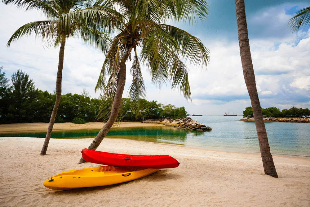 A yellow and red kayak on a sandy beach with palm trees and blue water in the background.