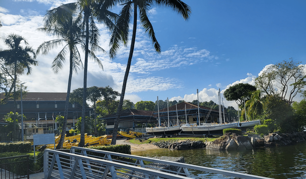 Colorful sailboats anchored at the waterfront of an outdoor adventure education center in Singapore, surrounded by lush greenery and a clear blue sky. 