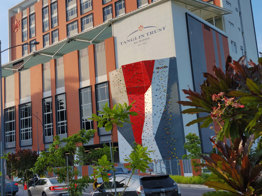 Photo of a colorful climbing wall at Tanglin Trust School in Singapore, featuring various climbing holds and routes. The Tanglin Trust School logo is prominently displayed above the wall.