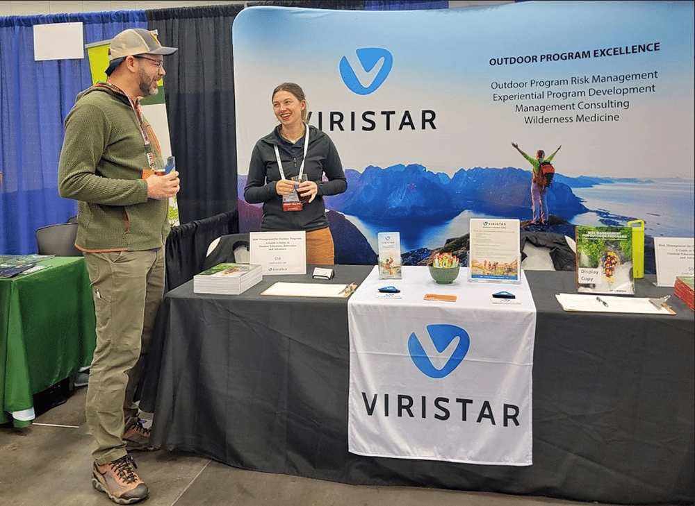 hoto of Viristar's exhibition table at a conference, featuring a Viristar team member engaging in conversation with a conference attendee. The table is adorned with promotional materials, brochures, and a banner showcasing Viristar's branding.