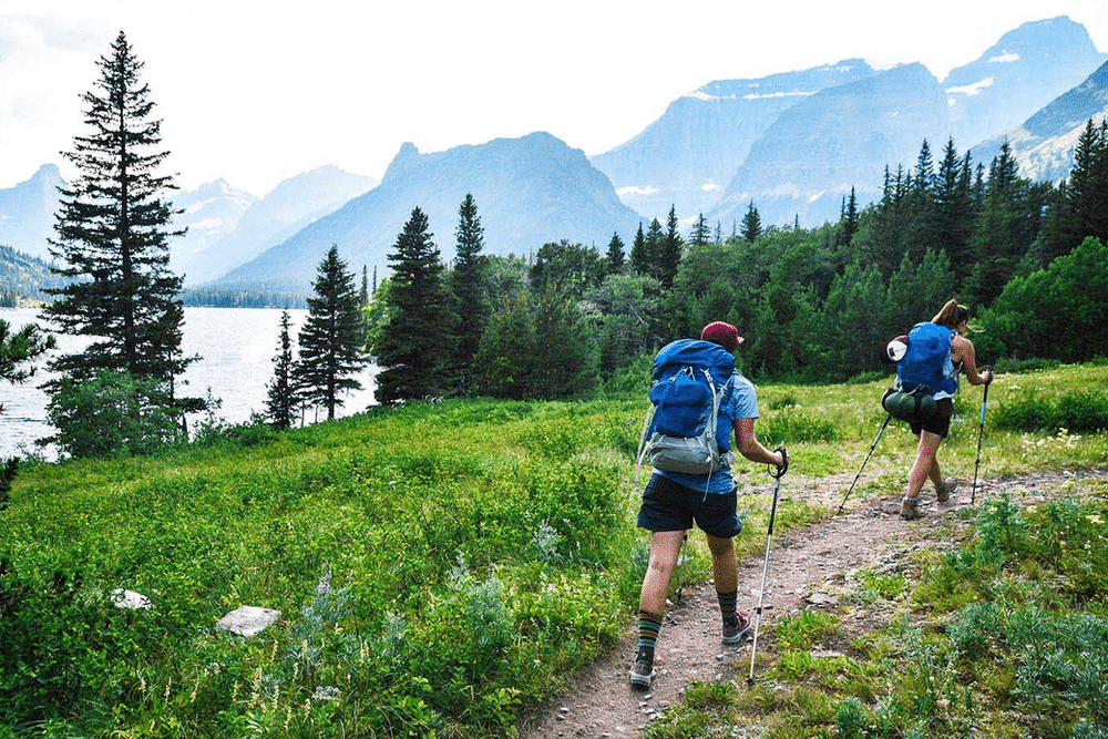 Two backpackers wearing blue backpacks and using trekking poles, hiking through a scenic, mountainous area.