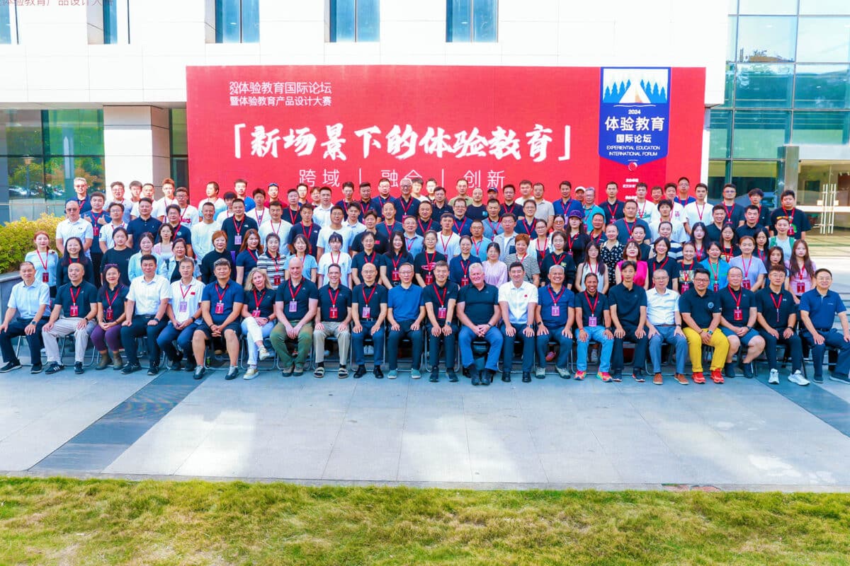 Group photo of participants at the Experiential Education International Forum. The diverse group stands together, smiling and posing in front of a backdrop that displays the event's logo. 