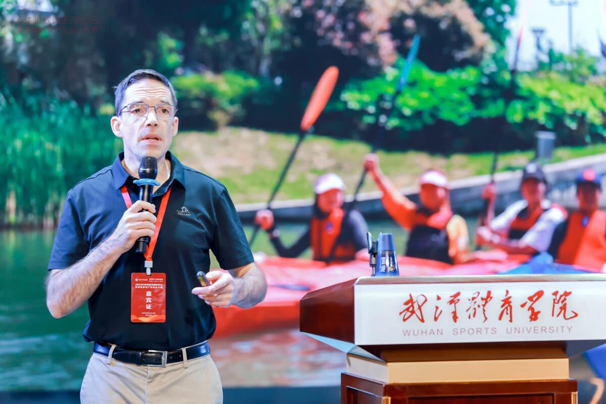 Viristar’s Director, Jeff Baierlein, presenting at a podium while wearing a dark blue shirt. He stands next to a large screen displaying a photo of people kayaking, enhancing his talk on outdoor safety and risk management. 