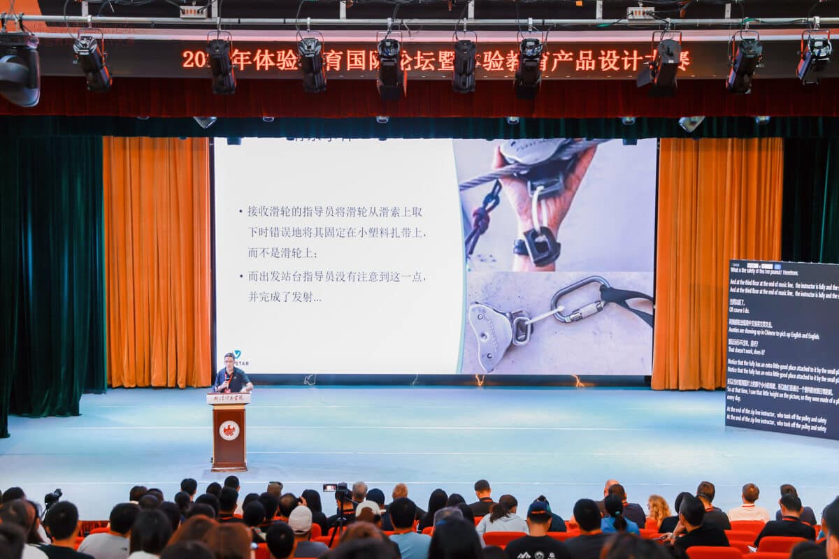 Viristar's Director stands on a large stage delivering a presentation to an engaged audience. The presentation screen behind him displays various safety-related images, emphasizing the importance of safety in outdoor and adventure programs.