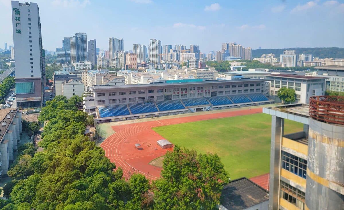 A collage showcasing Wuhan Sports University, featuring a vibrant green field at the center, surrounded by multi-tiered stadium seating. In the background, the city skyline of Wuhan is visible.