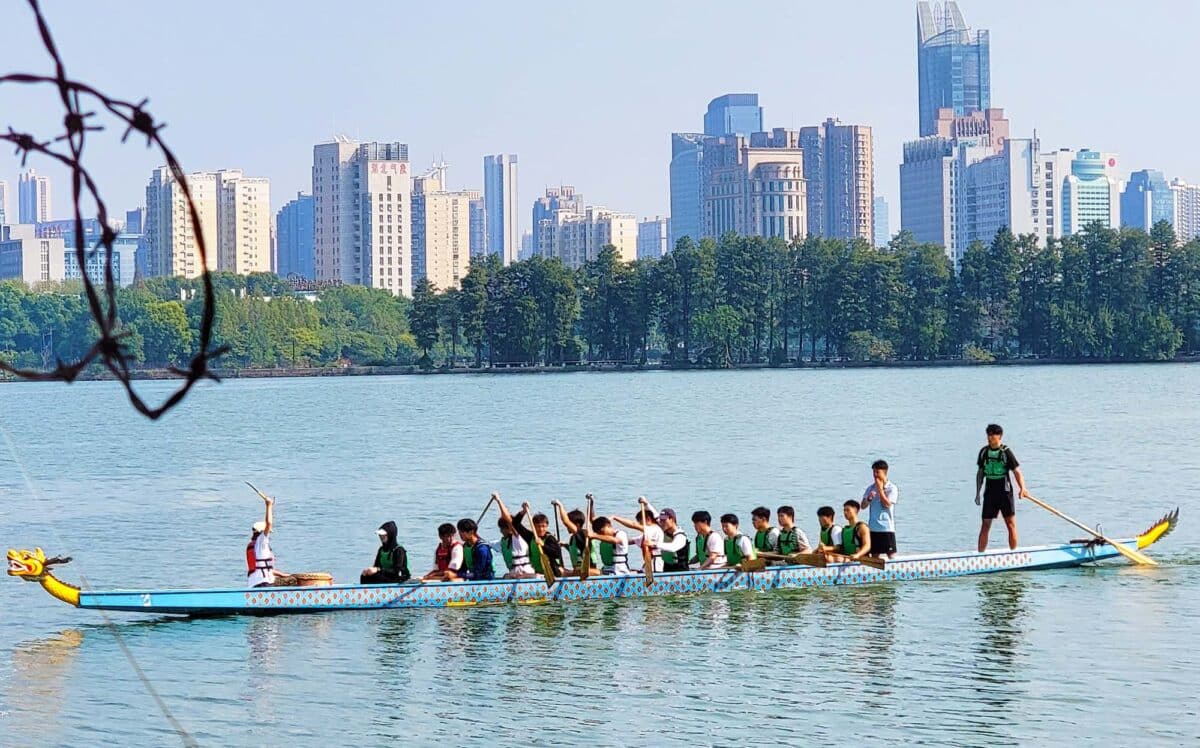 A group of students from Wuhan Sports University practicing dragon boating on Dong Lake in Wuhan. The scene captures a colorful dragon boat on the water, with students wearing life jackets, paddling in unison. Lush greenery and city buildings are in the distance.