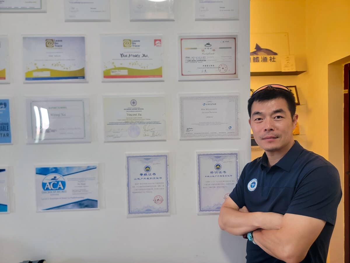 Dr. Xu stands proudly next to a wall displaying his impressive array of certifications from various international institutions, including Viristar, American Canoe Association, Leave No Trace, Association for Challenge Course Technology, and China Mountaineering Association, among others. The certifications showcase his expertise in outdoor education and risk management.