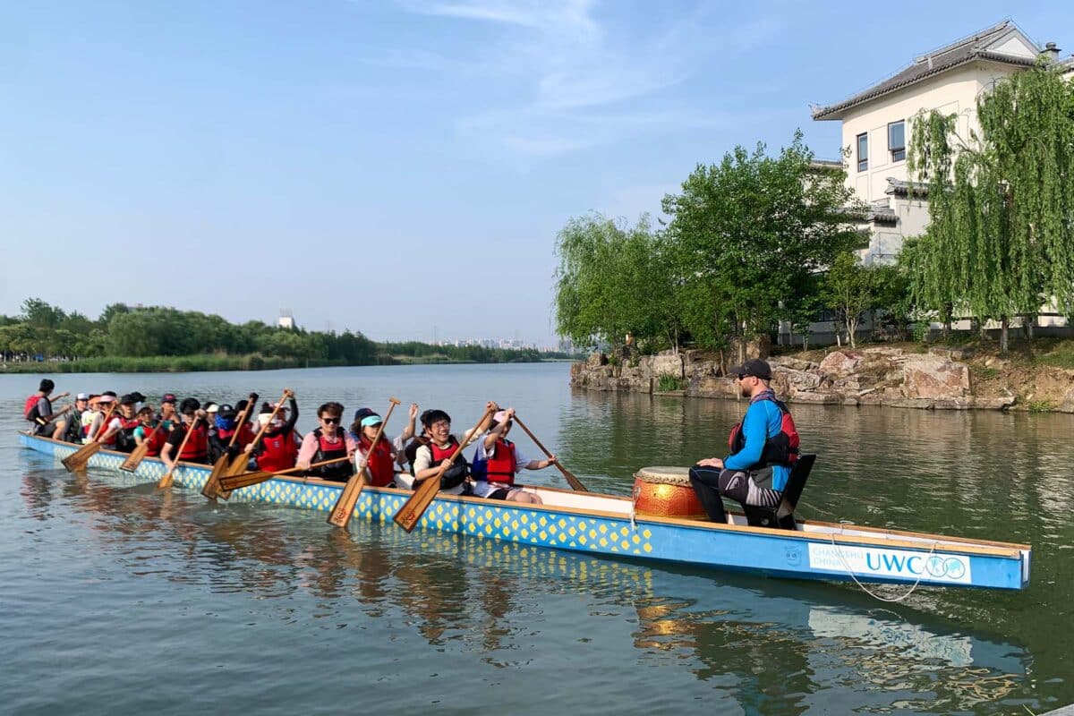 A group of people paddling together on a dragon boat in a calm lake. The team members, dressed in life vests, are actively rowing while a leader at the front sits next to a drum, guiding them. Trees and buildings are visible on the shore in the background.