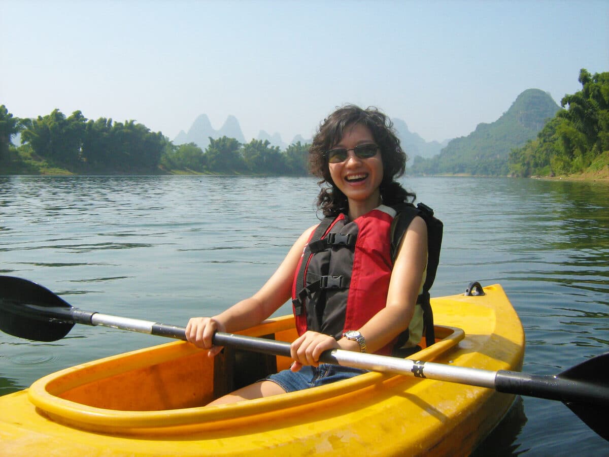 A woman wearing a red life vest paddles a yellow kayak on the tranquil waters of the Lijiang River, surrounded by lush green mountains and a clear blue sky.