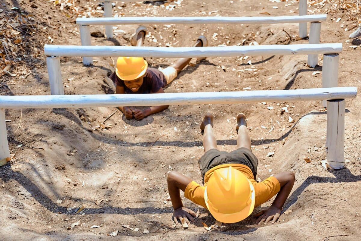 Participants at Lawrencedale Outdoors navigate an outdoor obstacle course, showcasing teamwork and determination as they crawl under wooden beams amidst a natural setting. 