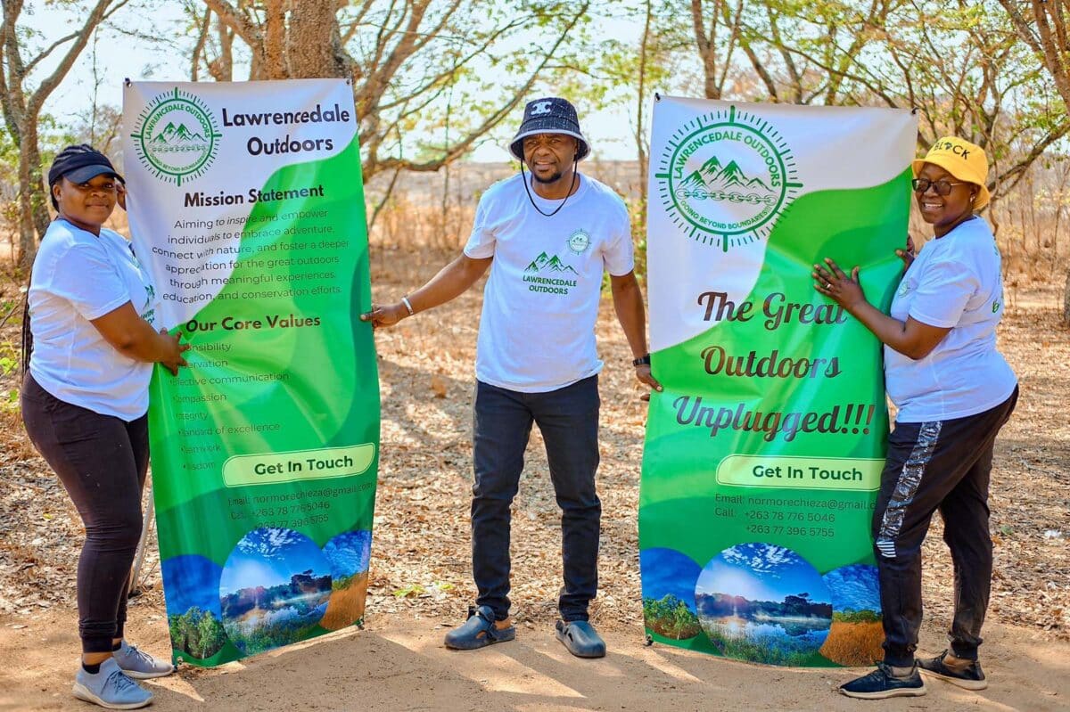 Three people wearing white *Lawrencedale Outdoors* t-shirts stand outdoors, holding two green and white banners for the organization. The banners display *Lawrencedale Outdoors'* mission statement and core values, emphasizing connection with nature and outdoor adventure. The setting is in a dry, wooded area, with trees and dry leaves in the background, and the group appears friendly and welcoming.