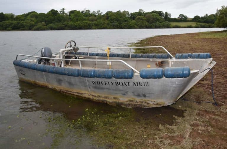 This image shows a Wheelyboat Mk III, an aluminum boat designed for accessibility, sitting partially on a grassy shore beside a body of water. The boat has blue fenders around the edges for protection and is equipped with a steering wheel and outboard motor. The background features lush green trees and rolling countryside under a cloudy sky.