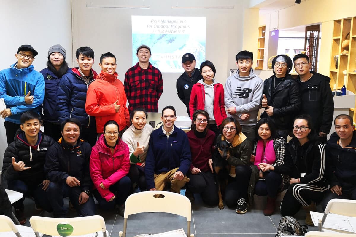 A diverse group of people, including men and women, pose together indoors for a group photo in front of a presentation screen that reads, "Risk Management for Outdoor Programs." They are dressed in warm clothing and some are smiling and giving thumbs-up gestures. 